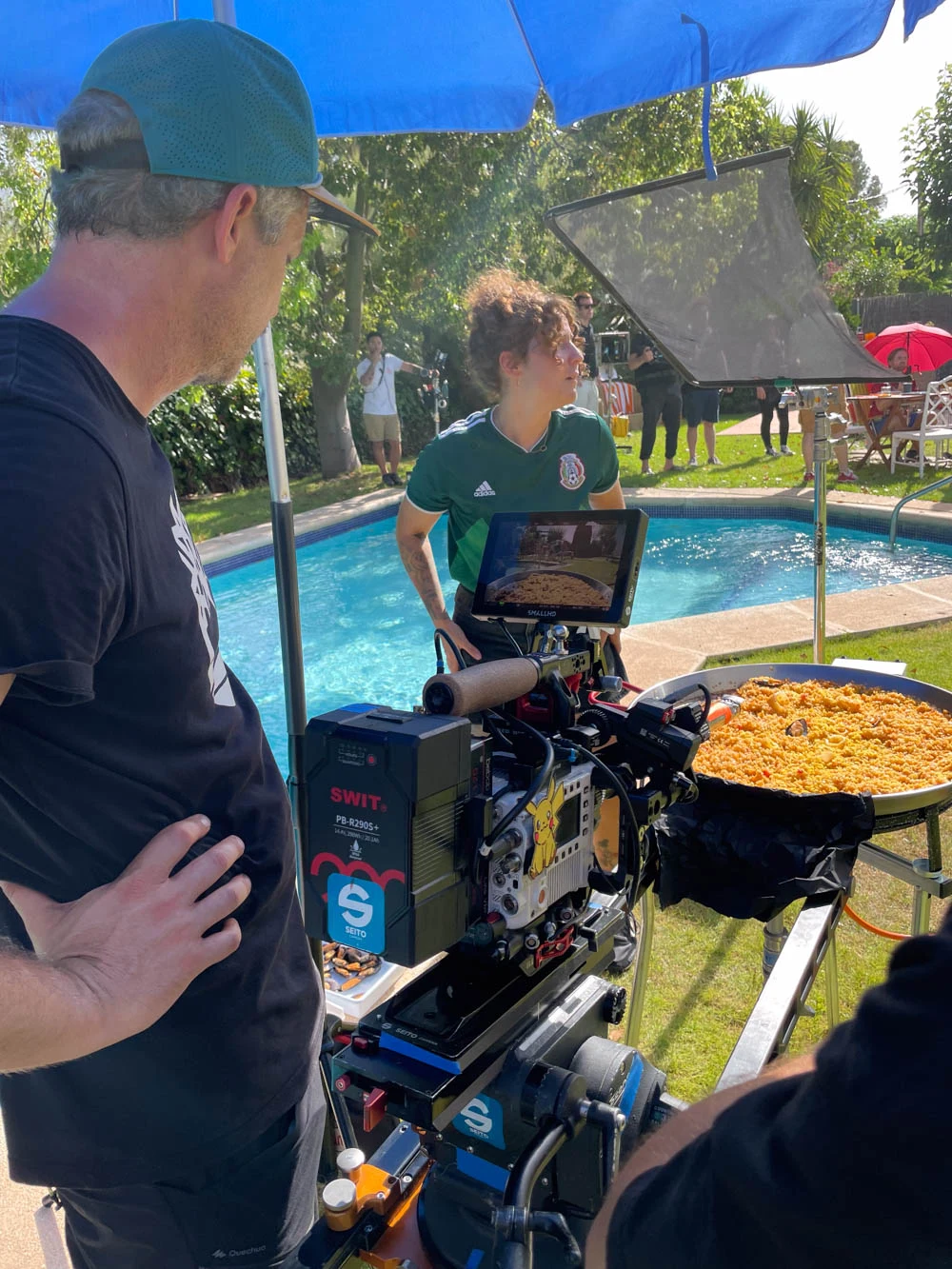 Camera framing a paella by a swimming pool, with Glassy Films crew and an actress on set